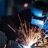 A worker wearing an industrial uniform and welding mask while working at a steel welding plant with sparks flying.