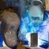 Two steel construction workers wearing protective gear and welding metal beams, with sparks flying in different directions.