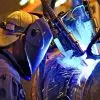A welder uses automated equipment on a large pipe. He wears protective gear and controls the machine with a remote.