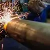 A welder works on a large steel pipe with equipment that sends sparks flying. He seems to be cutting or welding that section of the pipe.