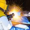 A technician operator welds a large metal tube structure in a factory workshop. They are wearing yellow protective gear.