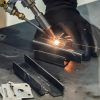 A professional welder uses a welding head for precise work on a small metal piece on a metal surface.