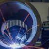 A welder with safety gear crouches inside a large metal pipe, performing a weld using a machine placed next to them.