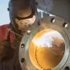 A welder in a protective mask and gloves carefully welds a steel pipe. Smoke emits at the contact point.