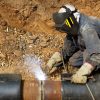 A welder works on a large metal pipe to secure the joint. They wear a mask and gloves for safety.