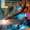 A welding robot with two arms operates on a steel beam. The machine works independently in a factory workspace.