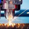 A laser CNC machine cutting through a thin metal sheet in a modern workspace. Blue lighting fills the environment.
