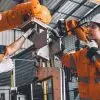 Two engineers in hard hats and orange jackets service a robot welding machine in an automated factory.