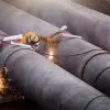 Three welders are assembling three large pipes at a construction site using professional equipment and safety gear.