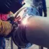 A welder stands close to a large metal pipe as they weld around it. They wear protective gear as they perform the weld.