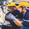 Two men in safety glasses and hard hats stand beneath a robotic arm with a welding torch as one holds a controller.